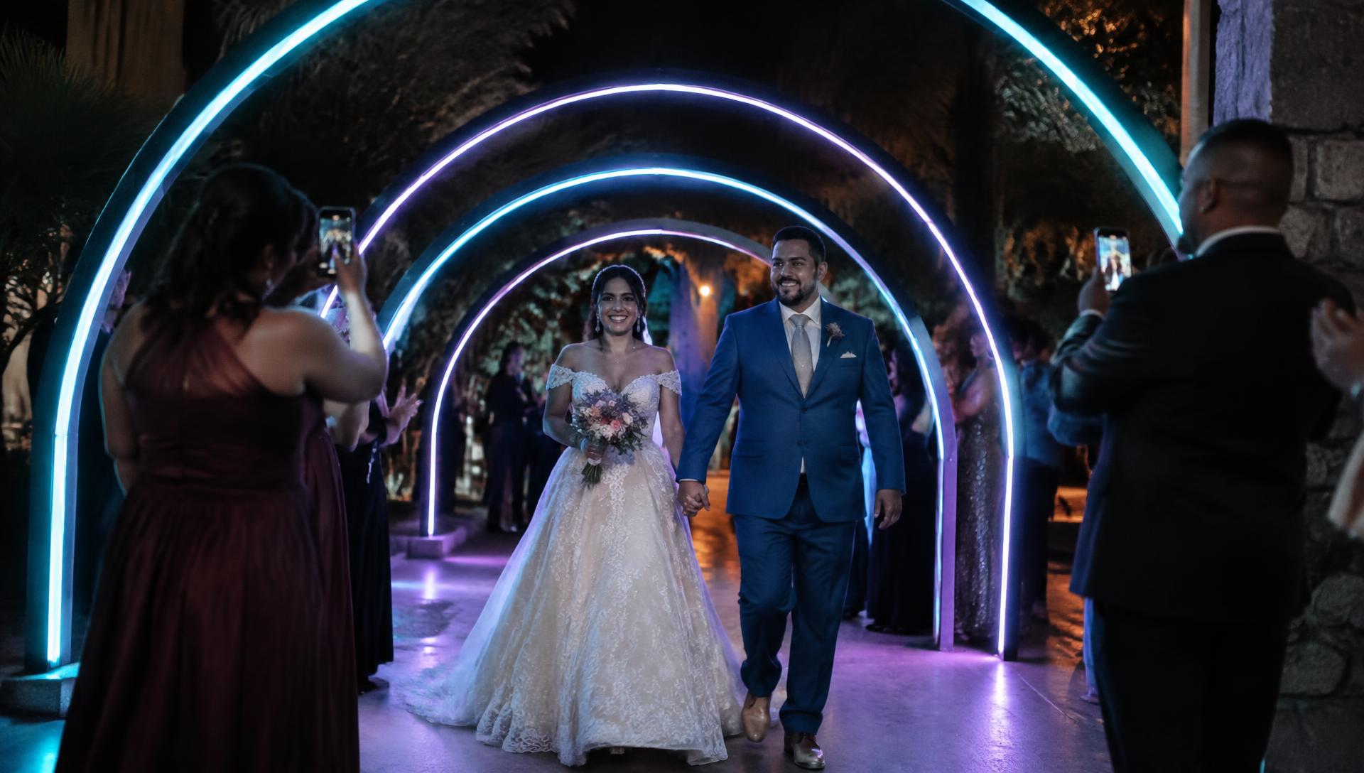 Bride and groom walking through a tunnel of glowing cyan and purple LED light archways as guests take photos